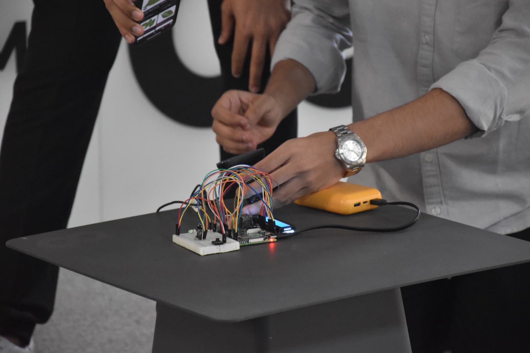 Hands setting up a breadboard prototype with jumper wires, a small board with a display and a power bank, while someone holds a phone showing the app.