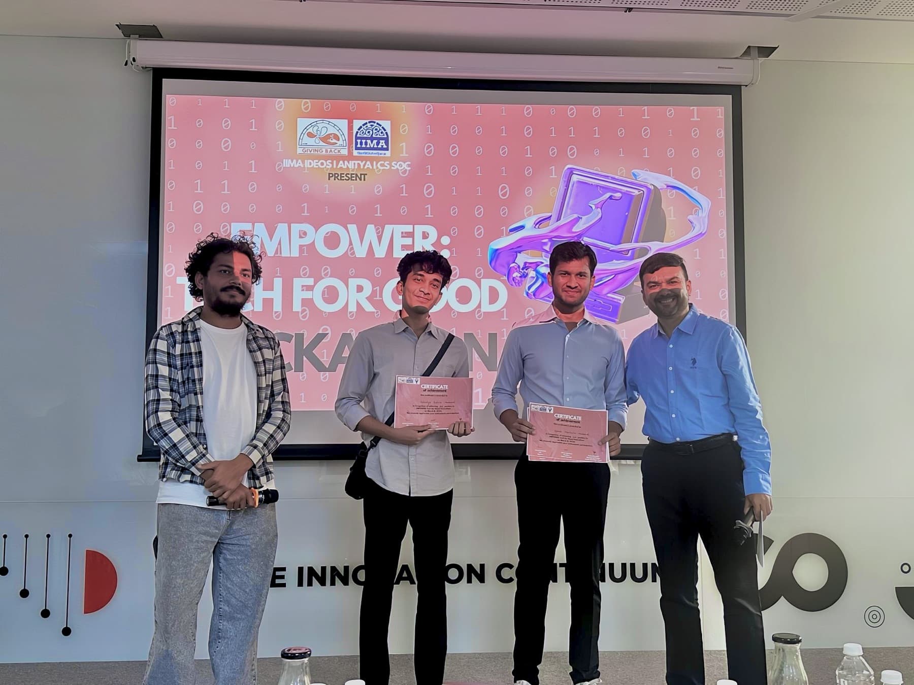 Vatsalya and a teammate holding certificates on stage in front of the EMPOWER: Tech for Good Hackathon screen, with two organizers beside them.